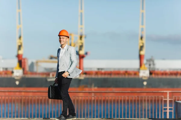 Handsome young unshaven successful business man in gray suit and protective construction orange helmet holding case, walking in sea port against cargo ship and crane background in summer time.