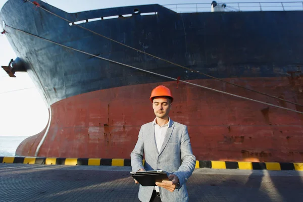 Handsome unshaven successful business man in gray suit, protective construction orange helmet holding tablet, standing in sea port against cargo rusty ship on water line background. Male with gadget