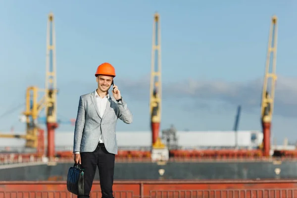 Handsome young unshaven successful business man in gray suit and protective construction orange helmet holding case, talking on mobile phone, walking in sea port against cargo ship, crane background