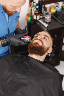 Erkek profesyonel kuaför hizmet istemci tarafından kesme makinesi. Zencefil acımasız şık yakışıklı genç kalın büyük sakal ve kısa saç Siyah pelerinli trendy saç kesimi almak. Işık beyaz Kuaför Salonu Oda