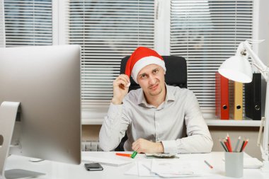 Masa başında oturan gömlek ve kırmızı Noel şapkalı yakışıklı üzgün ve kızgın iş adamı bilgisayar, yeni yılda modern monitör ile pencere arka plan üzerinde ışık Office belgeleriyle çalışma.