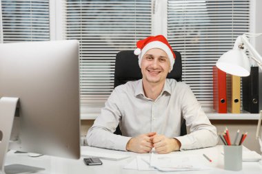 Işık Office belgeleri ile modern monitör bilgisayarda çalışan yakışıklı gülümseyen iş adamı gömlek ve masa başında oturan kırmızı Noel şapka kameraya pencere arka plan üzerinde seyir..