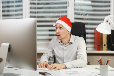 Yeni yıl bilgisayarda çalışan modern monitör, lamba, ışık Office belgeleri ile yakışıklı şok ve çapraşık iş adamı masa başında oturan gömlek, kırmızı Noel şapkalı klavyede eller