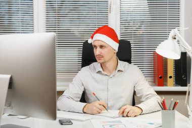 Yeni yıl bilgisayarda çalışan modern monitör, lamba, belge penceresi arka plan üzerinde ışık Office ile yakışıklı ciddi ve dalmış iş adamı masa başında oturan gömlek, kırmızı Noel şapkalı.