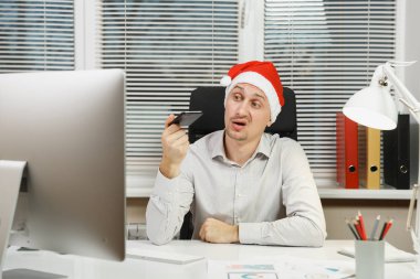 Yeni yıl bilgisayar, yakışıklı şok ve çapraşık iş adamı gömlekli, cep telefonu ile masaya oturan kırmızı Noel şapka çalıştığı modern monitör, lamba, ışık Office belgeleri ile.