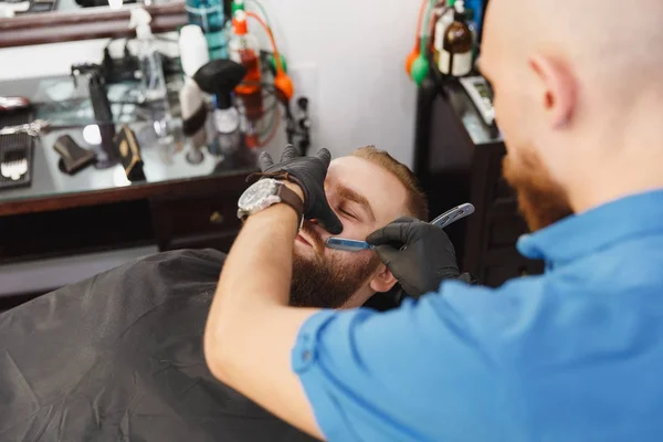 Male professional hairdresser serving client, shaving thick big beard straight razor. Ginger handsome stylish young man with short hair getting trendy haircut, black cape. Light white barber shop room