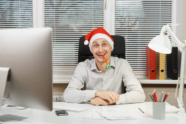 Hombre de negocios guapo sonriente en camisa y sombrero rojo de Navidad sentado en el escritorio, trabajando en la computadora con monitor moderno, lámpara, documentos y tubería de cumpleaños en la oficina de luz sobre fondo de ventana . — Foto de Stock