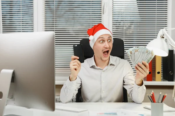 Yeni yıl ile modern sınıf başkanı bilgisayar, ışık office belgelerinde, üzgün, şok ve çapraşık iş adamı gömlekli, cep telefonu ve nakit para, Masa başında oturan kırmızı Noel şapka çalışma