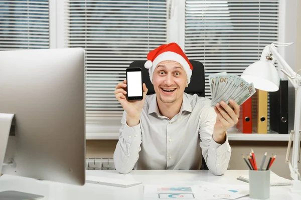 Sourire heureux homme d'affaires heureux en chemise, chapeau de Noël rouge assis sur le bureau avec téléphone portable et beaucoup d'argent comptant, travailler sur la nouvelle année à l'ordinateur avec moniteur moderne, documents dans le bureau léger . — Photo