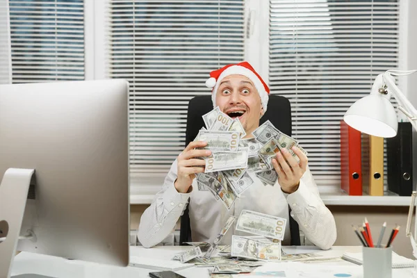Sorrindo feliz homem de negócios feliz na camisa, chapéu de Natal vermelho sentado na mesa com muito dinheiro em dinheiro, trabalhando no ano novo no computador com monitor moderno, lâmpada, pastas, documentos no escritório leve . — Fotografia de Stock