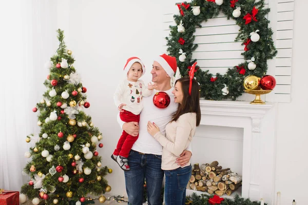 Szczęśliwych rodziców z cute synka w kapelusz. Chłopiec dziecko duży czerwony drzewo zabawka piłka stojący w urządzony nowy rok jasny pokój w domu na tle Boże Narodzenie wieniec. Rodzina, wakacje 2018 koncepcja. — Zdjęcie stockowe