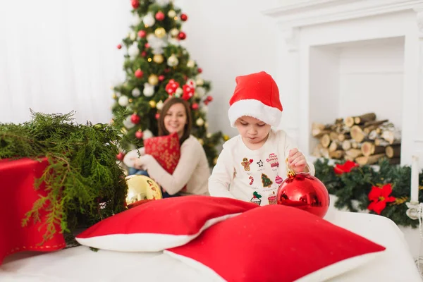 Happy young mother with cute little son in hat. Child boy with big red tree toy ball standing in decorated New Year light room at home. Christmas good mood. Family, love and holiday 2018 concept. — Stock Photo, Image