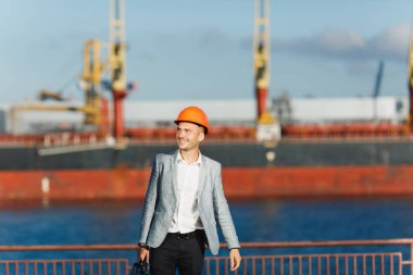 Gri takım elbise ve koruyucu inşaat turuncu yakışıklı genç tıraşsız başarılı iş adamı liman kargo gemisi karşı yürüyüş holding durumda, kask ve arka planda yaz saati vinç.