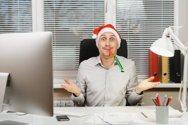 Bilgisayarda modern monitör, lamba, belgeler ve doğum günü boru pencere arka plan üzerinde ışık Office ile çalışan yakışıklı şok iş adamı masa başında oturan gömlek ve kırmızı Noel şapkalı.