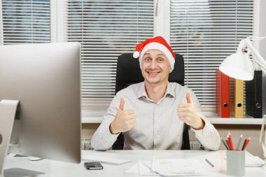 Yaşasın, çalışma ışık Office belgeleri ile modern İzleyicisi ile bilgisayar başında gösterilen yakışıklı gülümseyen iş adamı gömlek ve masa başında oturan kırmızı Noel şapka kameraya bakıyor.