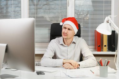 Masa başında oturan gömlek ve kırmızı Noel şapkalı yakışıklı üzgün ve kızgın iş adamı bilgisayar, yeni yılda modern monitör ile pencere arka plan üzerinde ışık Office belgeleriyle çalışma.