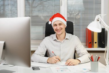Işık Office belgeleri ile modern monitör bilgisayarda çalışan yakışıklı gülümseyen iş adamı gömlek ve masa başında oturan kırmızı Noel şapka kameraya pencere arka plan üzerinde seyir..
