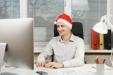 İş adamı gömlek ve masada oturan kırmızı Noel şapka gülümseyen yakışıklı, ışık Office belgeleri ile modern monitör ile bilgisayarda çalışma penceresi arka plan üzerinde klavye eller.