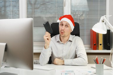 Yeni yıl bilgisayar, yakışıklı şok ve çapraşık iş adamı gömlekli, cep telefonu ile masaya oturan kırmızı Noel şapka çalıştığı modern monitör, lamba, ışık Office belgeleri ile.