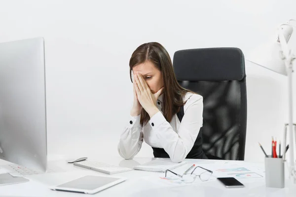 Tired perplexed and stress brown-hair business woman in suit sitting at the desk, covering her face with hands, working at contemporary computer with documents in light office on white background — Stock Photo, Image