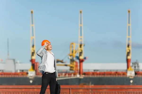 Handsome young unshaven successful business man in gray suit and protective construction orange helmet holding case, walking in sea port against cargo ship and crane background in summer time.