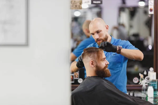 Male professional hairdresser serving client by scissors. Ginger handsome brutal stylish young man with thick big beard, short hair getting trendy haircut in black cape. Light white barber shop room. — Stock Photo, Image