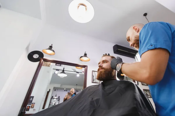 Męski profesjonalny salon fryzjerski klienta przez clipper. Ginger przystojny brutalny stylowe młody człowiek z brody duże grube i krótkie włosy coraz modne fryzury w czarnej pelerynie. Światło białe barber shop Pokój — Zdjęcie stockowe