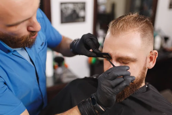 Cabeleireiro profissional masculino servindo cliente, tosquiando sobrancelhas com tesoura. Gengibre elegante jovem com barba grande grossa e cabelo curto ficando moderno corte de cabelo em capa preta. Sala de barbeiro leve — Fotografia de Stock