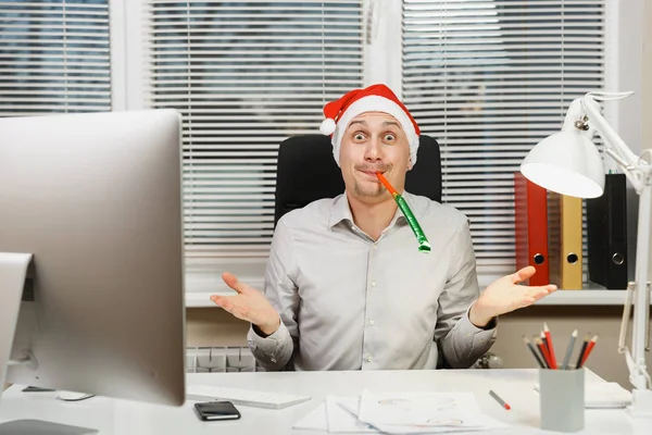 Bilgisayarda modern monitör, lamba, belgeler ve doğum günü boru pencere arka plan üzerinde ışık Office ile çalışan yakışıklı şok iş adamı masa başında oturan gömlek ve kırmızı Noel şapkalı.