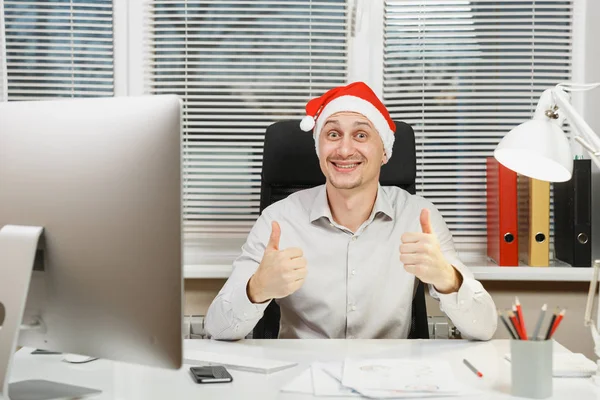 Yaşasın, çalışma ışık Office belgeleri ile modern İzleyicisi ile bilgisayar başında gösterilen yakışıklı gülümseyen iş adamı gömlek ve masa başında oturan kırmızı Noel şapka kameraya bakıyor.