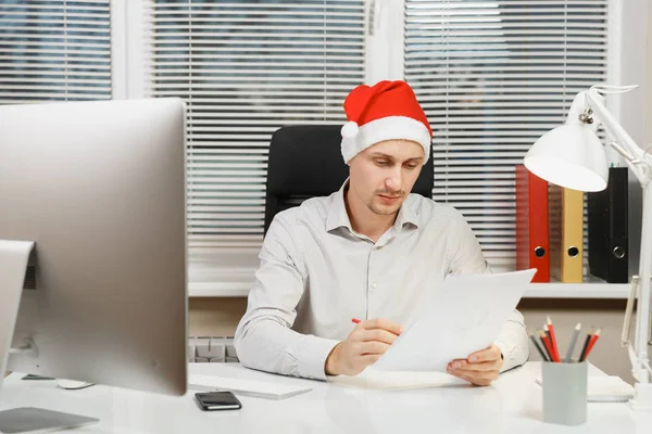 Guapo hombre de negocios serio y absorto en la camisa, sombrero rojo de Navidad sentado en el escritorio, trabajando en el nuevo año en el ordenador con monitor moderno, lámpara, documentos en la oficina de luz en el fondo de la ventana . — Foto de Stock