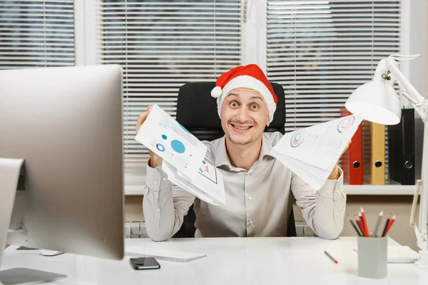 Guapo hombre de negocios sorprendido y perplejo en camisa, sombrero rojo de Navidad sentado en el escritorio, trabajando en el nuevo año en el ordenador con monitor moderno, lámpara, documentos en la oficina de luz en el fondo de la ventana . — Foto de Stock