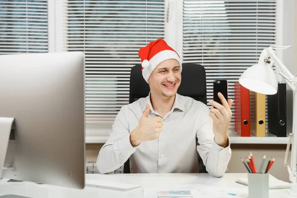 Hombre de negocios sonriente con camisa, sombrero rojo de Navidad sentado en el escritorio, haciendo selfie en el teléfono móvil, mostrando el pulgar hacia arriba, trabajando en el año nuevo en la computadora con monitor moderno, documentos en la oficina ligera . — Foto de Stock