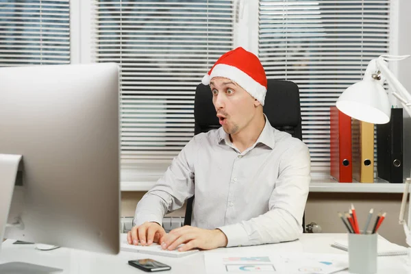 Yeni yıl bilgisayarda çalışan modern monitör, lamba, ışık Office belgeleri ile yakışıklı şok ve çapraşık iş adamı masa başında oturan gömlek, kırmızı Noel şapkalı klavyede eller