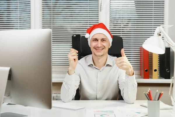 잘생긴 웃는 비즈니스 남자 셔츠와 레드 크리스마스 모자와 휴대 전화, 책상에 앉아에 현대 모니터 컴퓨터, 문서 창 배경에 빛 사무실에서 새 해에. — 스톡 사진