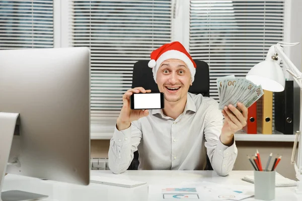 Sonriendo feliz hombre de negocios feliz en camisa, sombrero rojo de Navidad sentado en el escritorio con teléfono móvil y mucho dinero en efectivo, trabajando en el nuevo año en el ordenador con monitor moderno, documentos en la oficina de luz . — Foto de Stock