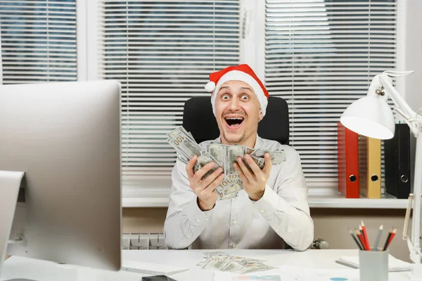 Sorrindo feliz homem de negócios feliz na camisa, chapéu de Natal vermelho sentado na mesa com muito dinheiro em dinheiro, trabalhando no ano novo no computador com monitor moderno, lâmpada, pastas, documentos no escritório leve . — Fotografia de Stock