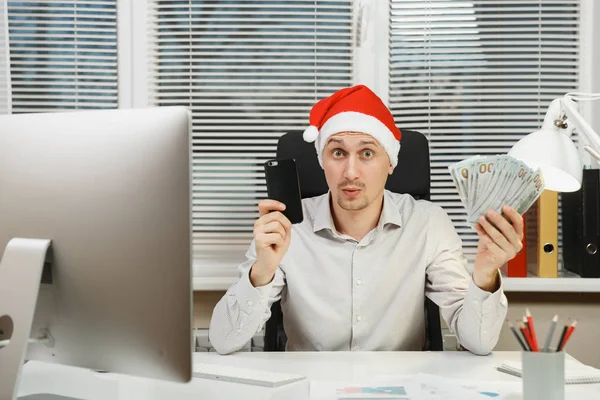 슬픈, 충격과 난처 한 사업가 셔츠, 휴대 전화 및 현금 돈, 책상에 앉아 빨간 크리스마스 모자에에서 현대 모니터 컴퓨터, 가벼운 오피스 문서에서 새 해에 노력 — 스톡 사진