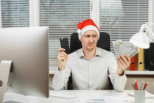Yeni yıl ile modern sınıf başkanı bilgisayar, ışık office belgelerinde, üzgün, şok ve çapraşık iş adamı gömlekli, cep telefonu ve nakit para, Masa başında oturan kırmızı Noel şapka çalışma