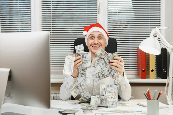 Sonriendo feliz hombre de negocios feliz en camisa, sombrero rojo de Navidad sentado en el escritorio con mucho dinero en efectivo, trabajando en el nuevo año en el ordenador con monitor moderno, lámpara, carpetas, documentos en la oficina de luz . —  Fotos de Stock