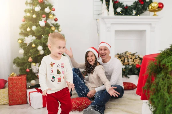 Feliz joven padres alegres con lindo hijo pequeño en sombrero rojo. Niño en la sala de luz en casa con árbol de Año Nuevo decorado y cajas de regalo. Navidad buen humor. Familia, amor y vacaciones 2018 concepto . — Foto de Stock