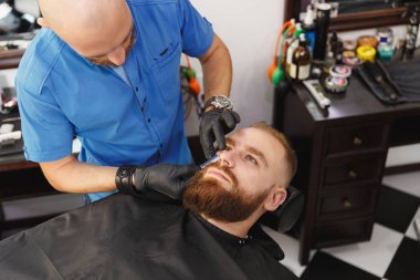 Erkek profesyonel kuaför hizmet istemci tarafından kesme makinesi. Zencefil acımasız şık yakışıklı genç kalın büyük sakal ve kısa saç Siyah pelerinli trendy saç kesimi almak. Işık beyaz Kuaför Salonu Oda