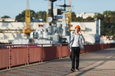 Yakışıklı genç tıraşsız başarılı iş adamı gri takım elbise ve durumda tutarak, cep telefonu üzerinde konuşurken, liman kargo gemisi, Vinç arka plan karşı yürüyüş koruyucu inşaat turuncu kask