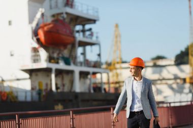 Gri takım elbise ve koruyucu inşaat turuncu yakışıklı genç tıraşsız başarılı iş adamı liman kargo gemisi karşı yürüyüş holding durumda, kask ve arka planda yaz saati vinç.