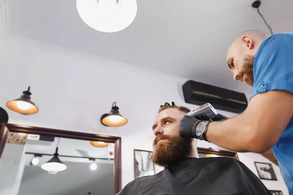 Cabeleireiro profissional masculino servindo cliente por clipper. Gengibre bonito jovem elegante brutal com barba grande grossa e cabelo curto ficando moderno corte de cabelo em capa preta. Sala de barbeiro branco claro — Fotografia de Stock