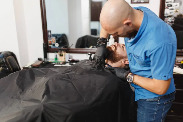 Чоловічий професійний перукар, що обслуговує клієнта, голиться товстою великою бородою прямої бритви. Імбир красивий стильний молодий чоловік з коротким волоссям стає модною стрижкою, чорна накидка. Світло-білий перукарня — стокове фото