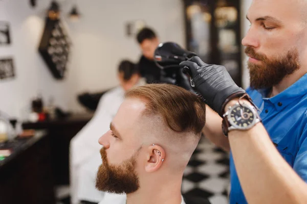 Male professional hairdresser serving client, drying hair with hairdryer. Ginger handsome brutal stylish young man with thick big beard and short hair getting trendy haircut. Light white barber shop. — Stock Photo, Image