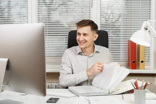 Красивий усміхнений бізнесмен у сорочці, що сидить за столом, працює за комп'ютером, чорною текою, документами у світлому офісі, дивиться на сучасний монітор, на фоні вікна. Менеджер або працівник . — стокове фото