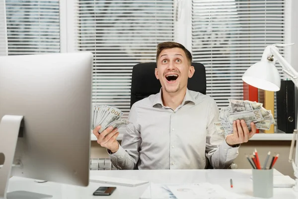 Buon uomo d'affari sorridente in camicia seduto alla scrivania con un sacco di soldi in contanti, lavorando al computer con monitor moderno, documenti in ufficio leggero sullo sfondo della finestra. Vincitore di manager o lavoratori . — Foto Stock