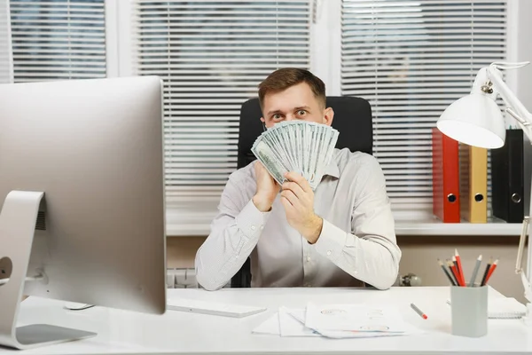 Handsome business man in shirt sitting at the desk with mobile phone hiding behind lot of cash money, working at computer with modern monitor, lamp, documents in light office. Manager or worker.